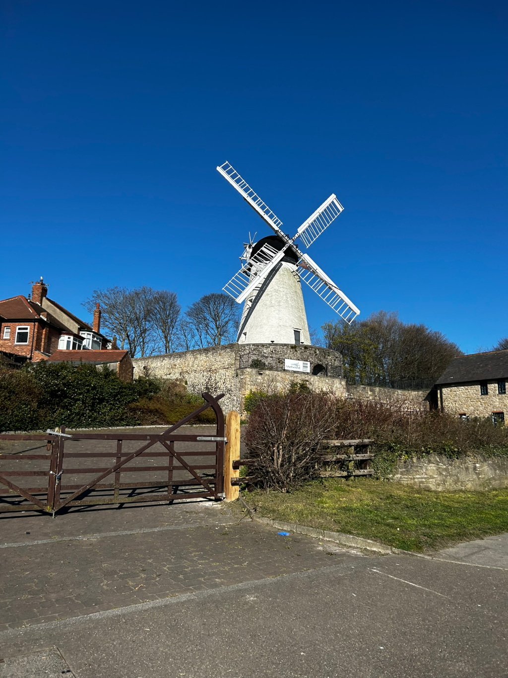 Windmill standing prominently against a clear blue sky. It's a white, tower mill with dark sails. The windmill is situated on a small hill, surrounded by a low stone wall. In the foreground is a wooden gate and some landscaping. Residential buildings are partially visible in the background to the left. The overall impression is one of a peaceful, rural scene.