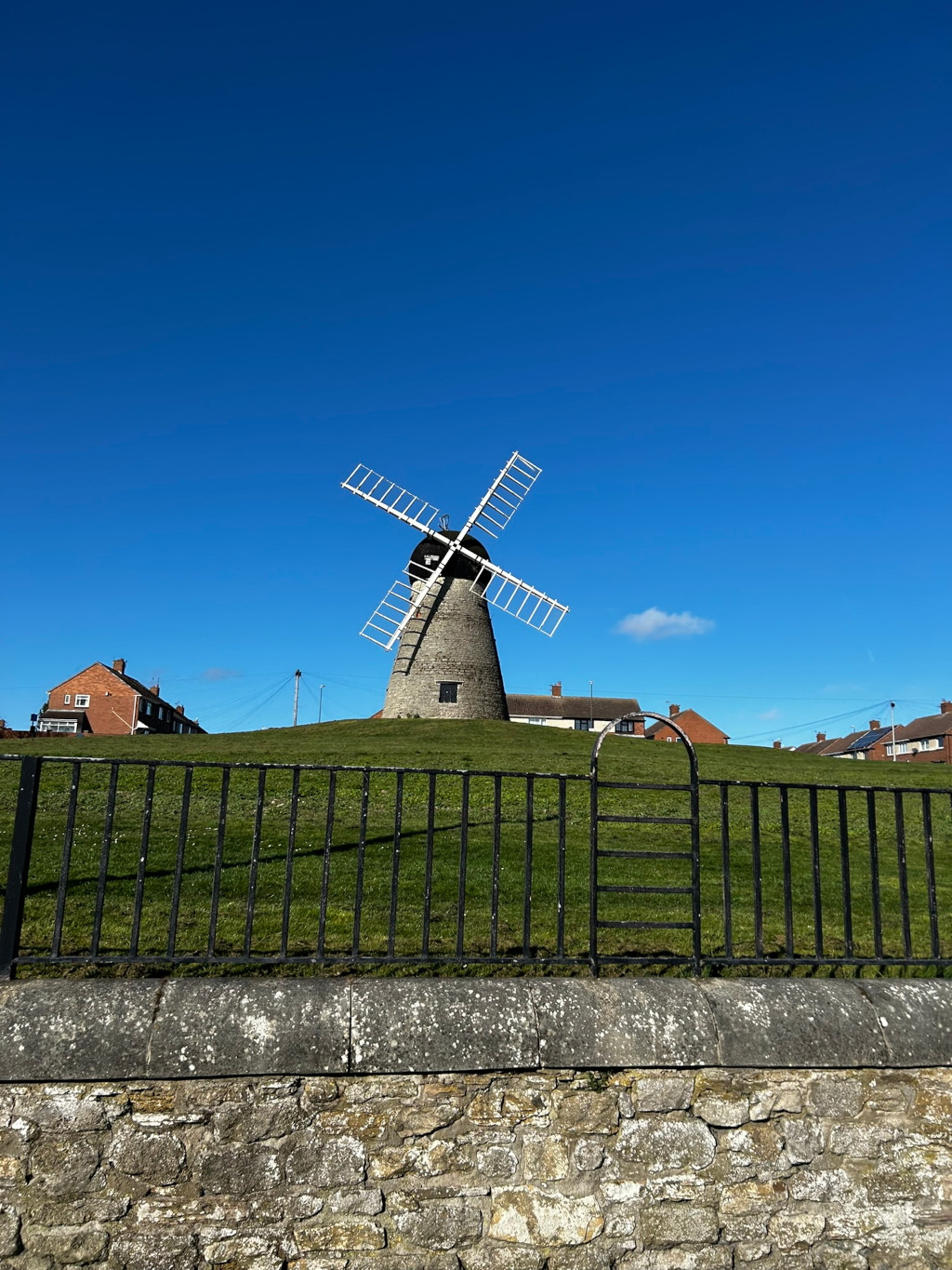 A stone windmill with white sails set against a clear blue sky.  The windmill sits atop a grassy hill, behind a black metal fence and a low stone wall. Residential houses can be seen in the background. The overall impression is one of a peaceful, idyllic setting.