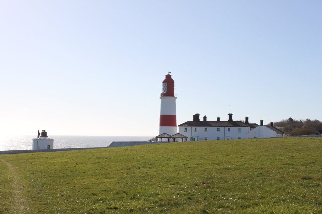 Coastal scene on a bright, sunny day. In the foreground is a vibrant green grassy field. In the mid-ground, there's a white building with multiple chimneys of a lighthouse keeper's house. Beyond that is the prominent, tall lighthouse, striped red and white. The sea is calm and visible in the background under a clear pale blue sky.