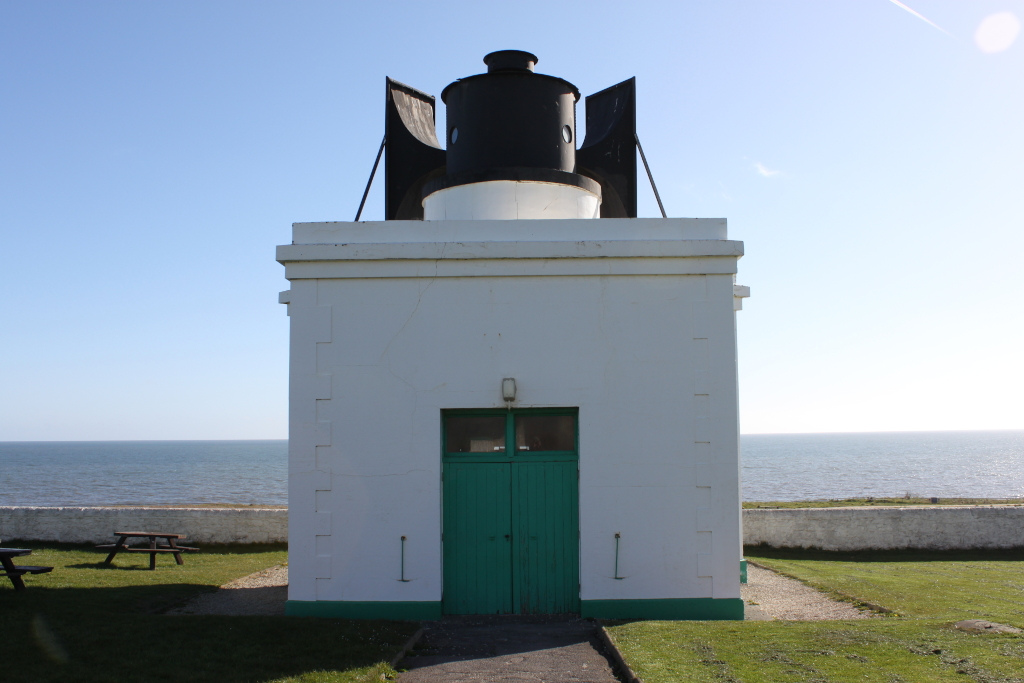 White, square building with a teal door, situated on a grassy area overlooking a calm sea under a clear blue sky. Two picnic tables are visible in the foreground. The top of the structure features a black, metallic fog signal. The overall impression is one of peaceful coastal serenity.