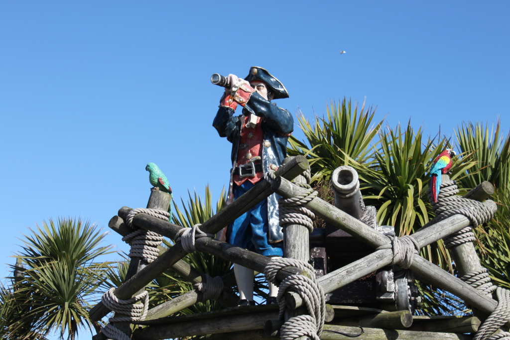 Statue of a pirate captain using a spyglass, perched atop a wooden structure reminiscent of a ship's crow's nest. Two parrots, one green and one red/yellow/blue, are also positioned on the structure. The backdrop is a clear blue sky and several palm-like plants.