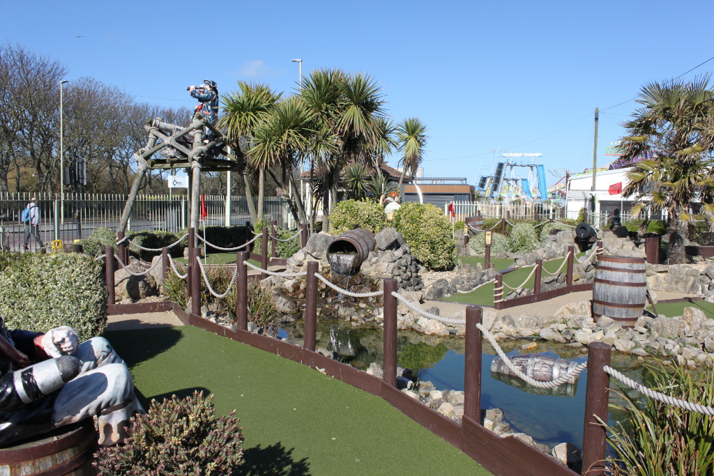 Pirate-themed mini-golf course. The foreground features artificial turf, rock formations, small ponds, wooden fences and rope railings. Pirate figureheads are visible, and there are barrels incorporated into the course design. In the background, a section of a seaside amusement park is partially visible. The overall scene suggests a bright, sunny day.
