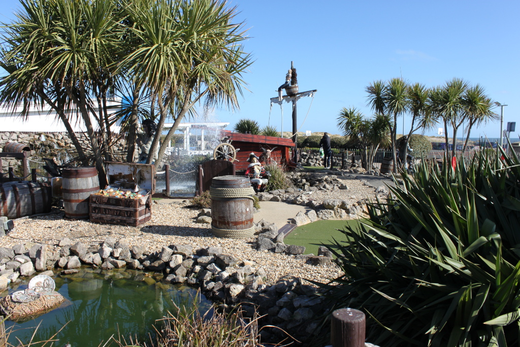 Pirate-themed section of a miniature golf course. The scene features a small artificial pond, rocks, numerous palm-like plants, wooden barrels, and a treasure chest. A small pirate ship replica is also visible, along with several pirate figures. In the background, there’s a glimpse of a building and other parts of the course. The overall setting and styling suggest a sunny, outdoor location designed for entertainment and recreation.