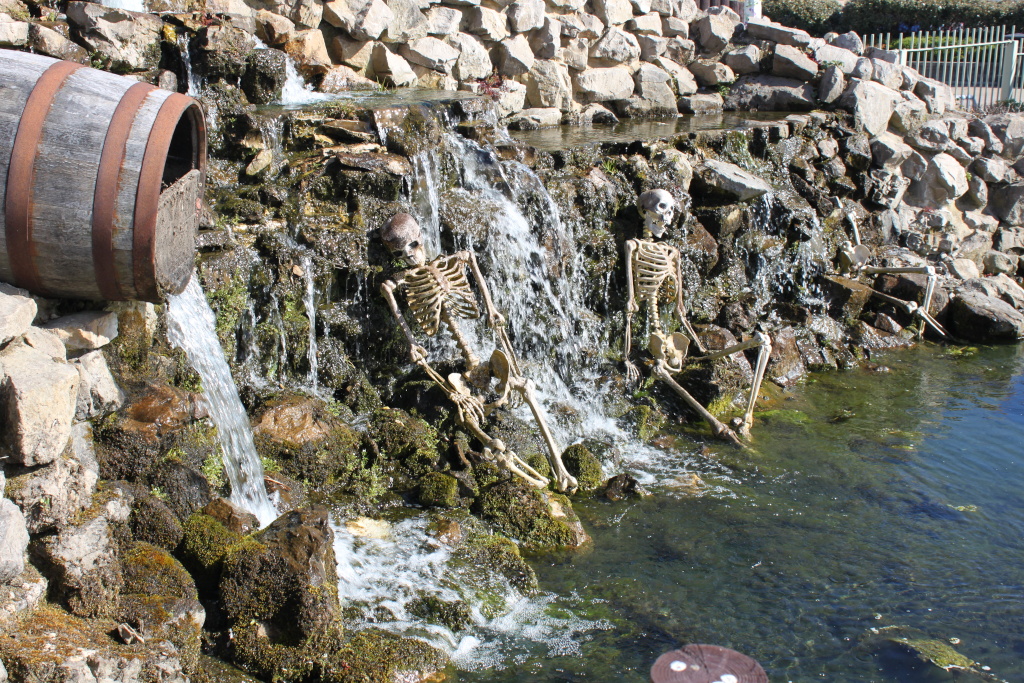 Three skeletons sitting in a small waterfall. A wooden barrel is situated at the top of the waterfall, from which water flows down the rocks. The scene is set in a small pond with mossy rocks and a stone wall in the background.