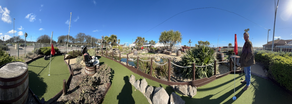 Panoramic image shows an outdoor pirate-themed miniature golf course on a sunny day. The course features artificial turf, wooden barrels, pirate figures, small ponds, and rock formations. Charlie stands near a putting green, seemingly ready to take a shot. The overall atmosphere is bright, cheerful, and family-friendly.