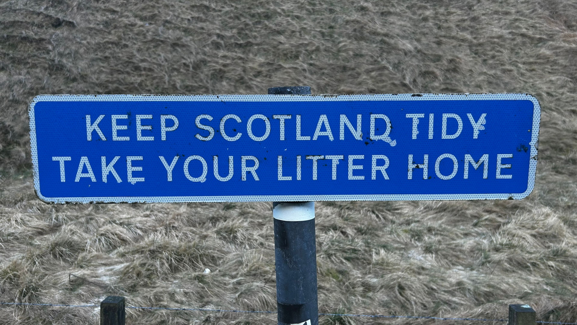 A blue sign with white lettering that reads KEEP SCOTLAND TIDY TAKE YOUR LITTER HOME. The sign is mounted on a dark post in front of a background of dry, light brown grass or moorland. The sign shows some wear and tear.
