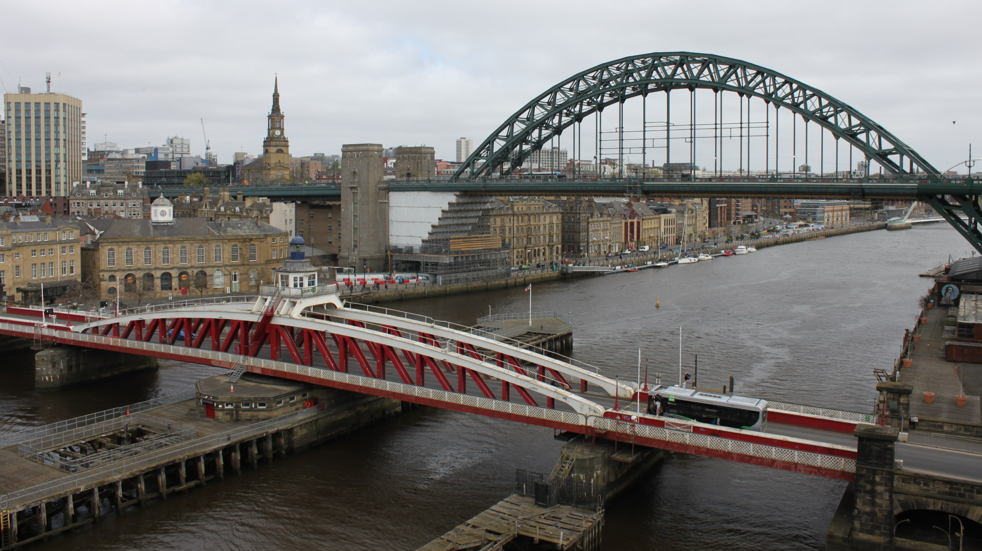 High-angle view of the Tyne River in Newcastle upon Tyne, England. The foreground features a red swing bridge, with a bus crossing it. In the middle ground, the river flows, with various boats visible.  The background includes the iconic Tyne Bridge (a steel arch bridge), numerous buildings, and a church steeple. The overall scene depicts a blend of industrial architecture and river traffic against a cloudy sky.