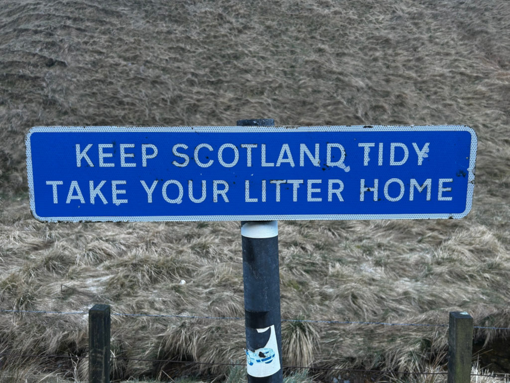 Blue sign with white lettering that reads KEEP SCOTLAND TIDY TAKE YOUR LITTER HOME. The sign is mounted on a dark post in front of a background of dry, light brown grass or moorland. The sign shows some wear and tear.