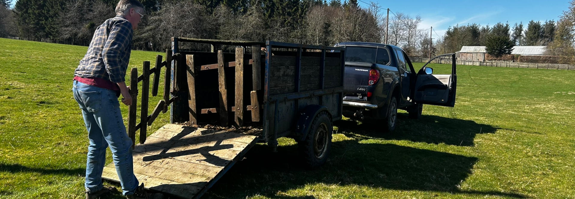 Charlie loading sections of a wooden fence into a trailer attached to a dark-coloured pickup truck. The setting appears to be a rural or agricultural area with a grassy field and trees in the background. The man is wearing a plaid shirt and jeans.