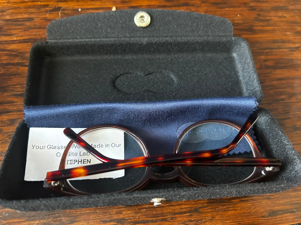 Pair of tortoiseshell-framed glasses resting in their dark-coloured hard case. A small card is visible inside the case, partially obscuring the glasses, indicating that the glasses were made on-site. The overall impression is one of new, carefully packaged eye wear.