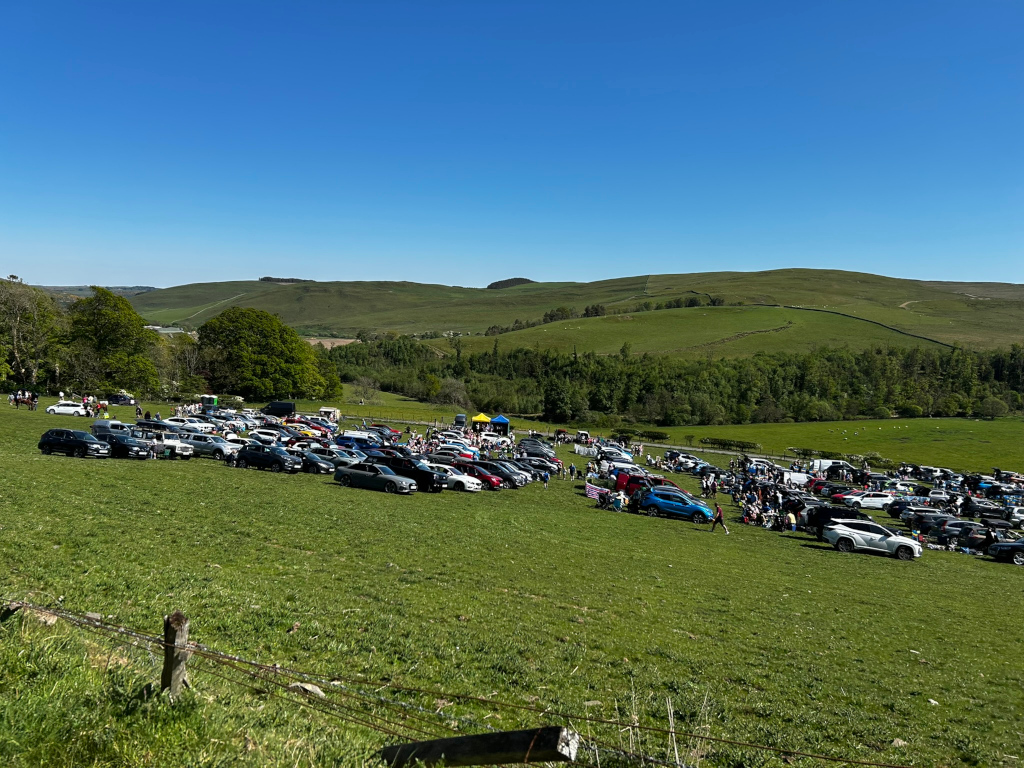 Large gathering of people and cars in a grassy field, set against the backdrop of rolling green hills under a clear blue sky. The cars are parked in a somewhat haphazard but largely organised manner, suggesting an outdoor event or gathering of some kind. The scene is peaceful and sunny, indicating a pleasant day.