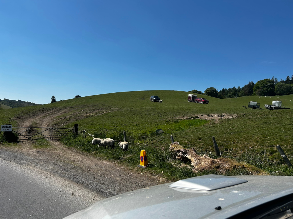 Grassy hillside on a sunny day.  In the middle ground, there are a couple of vehicles, one appearing to be a horse trailer, and a few sheep grazing on the hill. There's a car park sign visible near the bottom left, indicating what appears to be a rural or countryside location. A road or track leads towards the hill's base. The foreground shows a part of a car's roof and windshield, suggesting that the photo was taken from inside a vehicle. The overall scene is peaceful and pastoral.