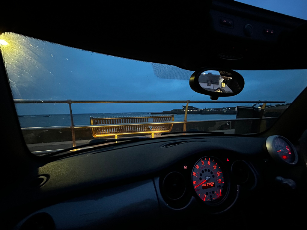 Interior view from the driver's seat of a car parked near a seaside. The focus is on the view through the windshield, which depicts a calm, dark-blue sea under a twilight sky. A park bench sits on the seawall in the foreground, with a hint of a distant town or harbour visible. The car's dashboard and rear view mirror are partially visible, adding to the overall tranquil and slightly melancholic mood.