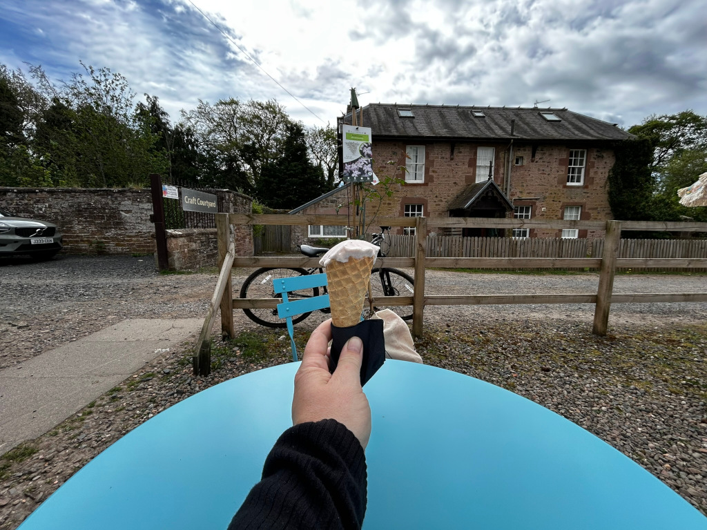 Leonie's hand holding an ice cream cone at a table outside a stone building. The building looks like a converted house or small business, perhaps a craft centre, as indicated by the Craft Courtyard sign. A bicycle is visible in the background, and the overall setting appears to be a peaceful, possibly rural location on a somewhat cloudy day. The scene suggests a moment of simple pleasure and relaxation.