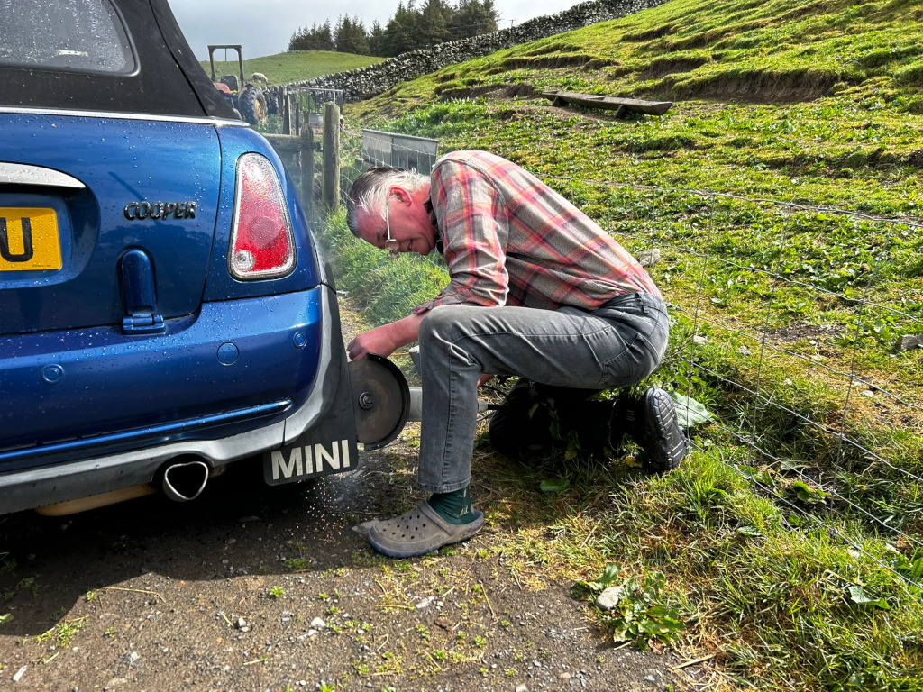 Charlie crouching beside a blue Mini Cooper, using an angle grinder to cut off part of the car's rear bumper. The setting appears to be a rural area with a grassy field and a fence in the background. He is wearing a plaid shirt and jeans. Sparks are visible from the grinder, indicating metal is being cut.