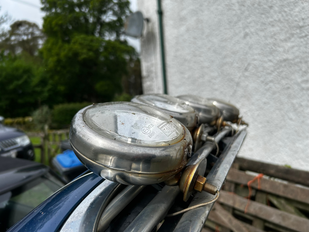 Close-up view of three vintage, circular, chrome-plated driving lamps mounted on a vehicle's roof rack. The lamps appear to be older and show signs of wear and age, including some surface discoloration and rust. The background is blurred but shows a residential setting with green foliage and a portion of a building’s wall. The focus is sharply on the lamps and their mounting hardware.