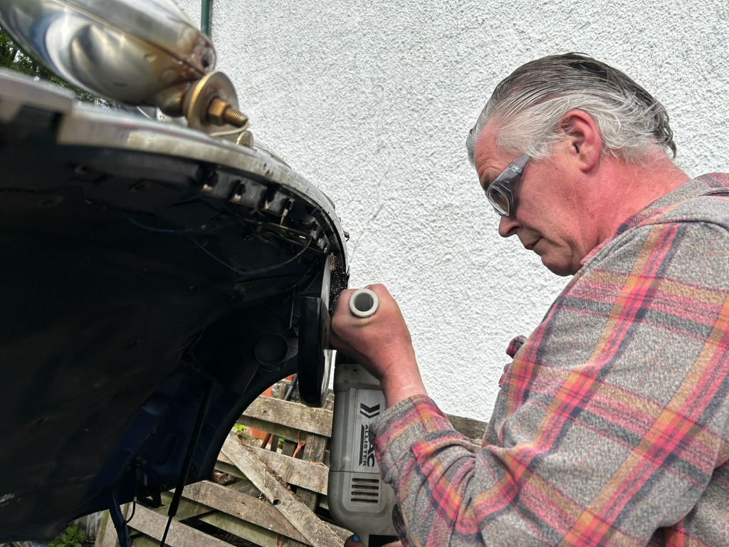 Charlie using a power tool to smooth the edge of a car's body panel. He's wearing safety glasses and a plaid shirt, working outdoors against a light-colored wall. The focus is on the man and the detail work he is performing on the vehicle.