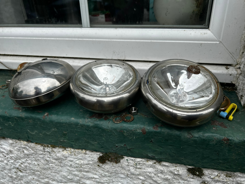 Three vintage or antique car headlights or spotlights resting on a windowsill. They appear to be made of metal with glass lenses, showing signs of age and wear. A few small metal parts and a couple of small pieces of debris are also visible near them on the windowsill.
