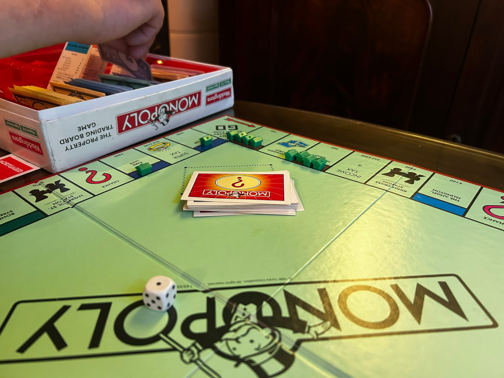 Game of Monopoly in progress. Charlies hand is seen placing money into the game's box. The board is partially visible, showing several properties with houses and Chance cards. The overall impression is one of casual, perhaps friendly, competition. The focus is on the game itself, implying leisure and possibly social interaction. There is no deeper moral or story explicitly shown, but the game itself is symbolic of competition, risk, and the pursuit of wealth.