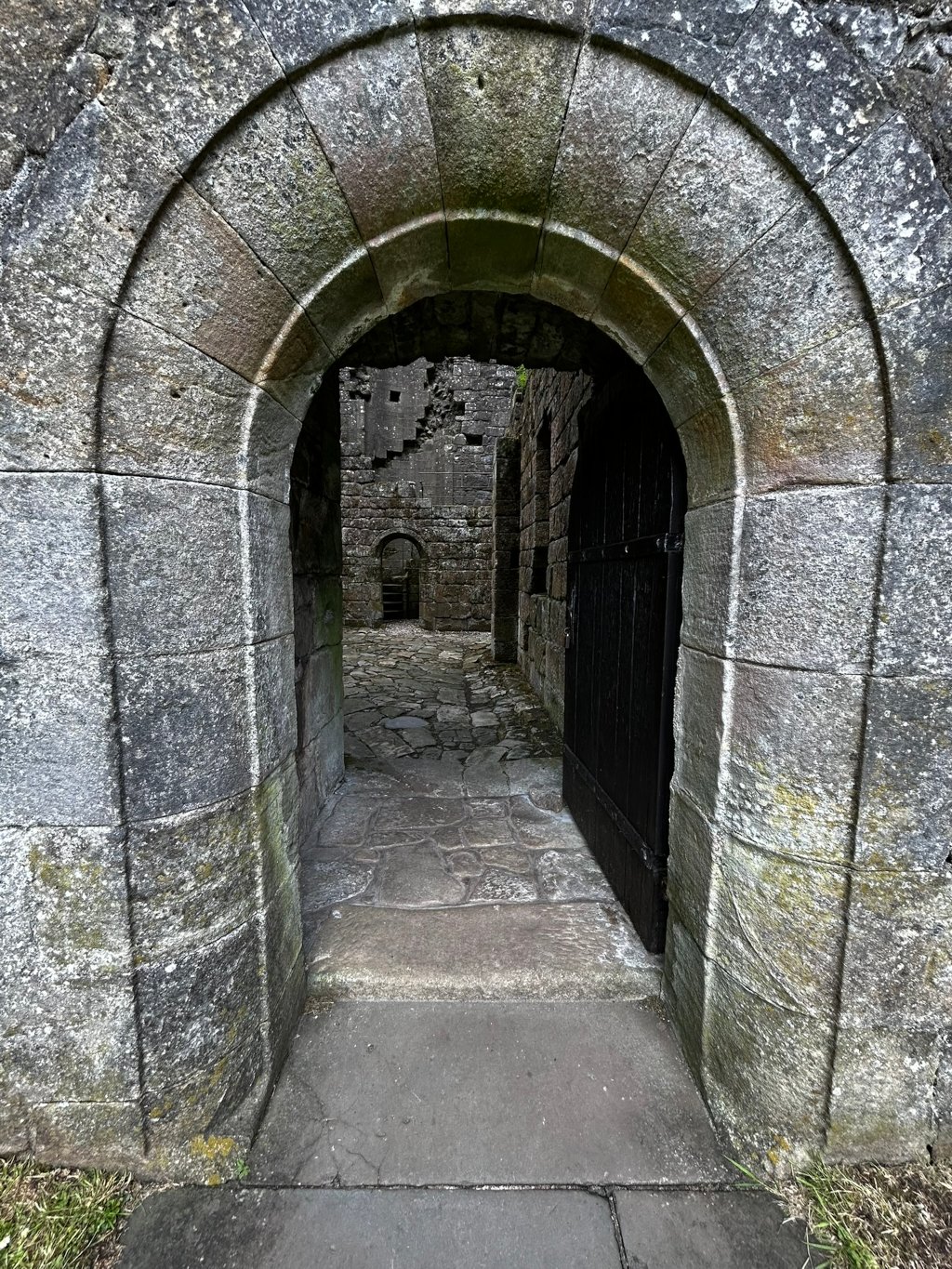 Stone archway, seemingly part of a ruin or ancient structure. The arch is made of weathered grey stone, showing signs of age and moss growth. Beyond the archway, a dark wooden door is ajar, revealing a paved pathway leading further into a stone courtyard or building, also appearing to be in ruins.