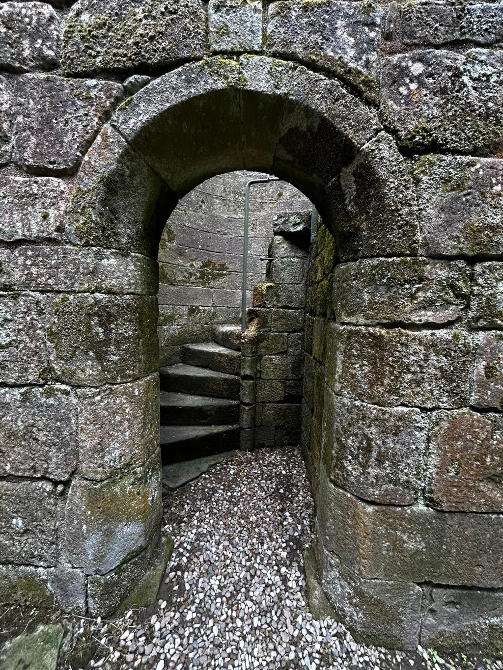 Stone archway leading to a spiral staircase within a stone structure, possibly a ruin or ancient building. The stones are weathered and covered in moss, suggesting age and exposure to the elements. A simple metal railing is visible on the staircase. The ground at the base of the archway is covered in small, light-coloured gravel.