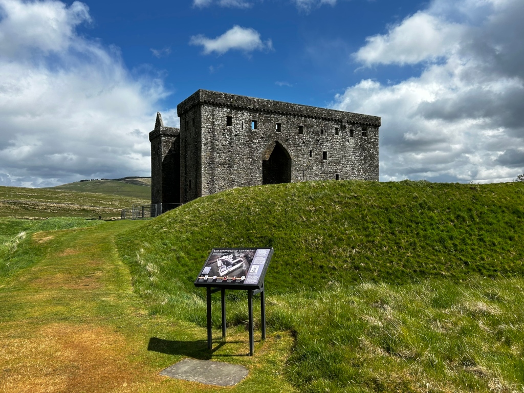 Ruins of a stone castle, specifically the keep, sitting atop a grassy mound. A modern informational sign stands in the foreground, providing details about the site. The background features a gently rolling, grassy landscape under a partly cloudy sky.