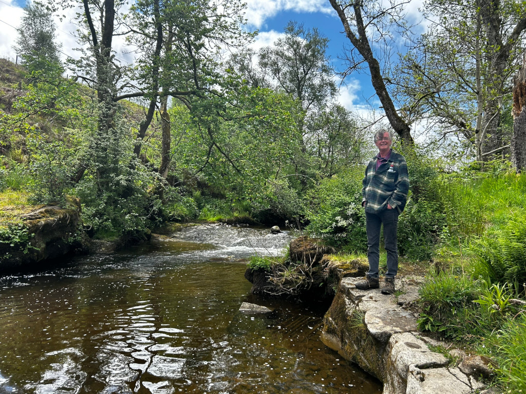 Charlie standing on a rock next to a small river. The river flows through lush green vegetation, and the overall setting appears to be a tranquil, natural environment. Charlie is casually dressed and appears to be enjoying the scenery. The sky is partly cloudy.