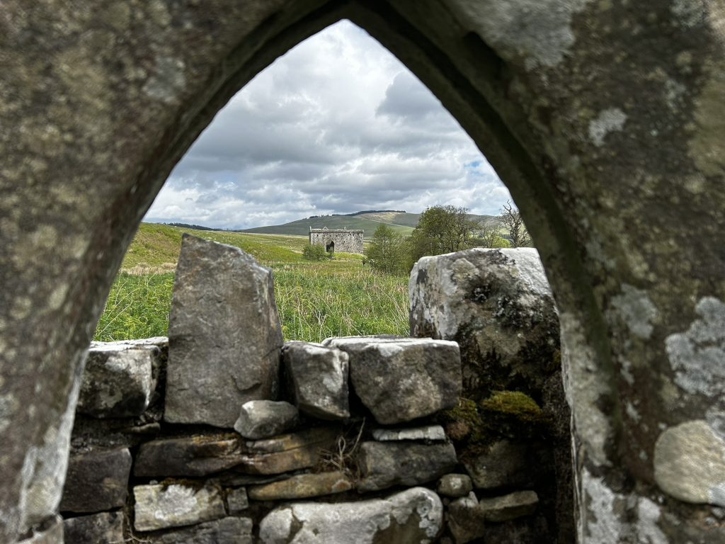 View through an arched opening in a weathered stone wall. Beyond the wall, a grassy field stretches towards a distant, small stone structure, possibly a ruin or tower, set against a backdrop of rolling hills under a cloudy sky.