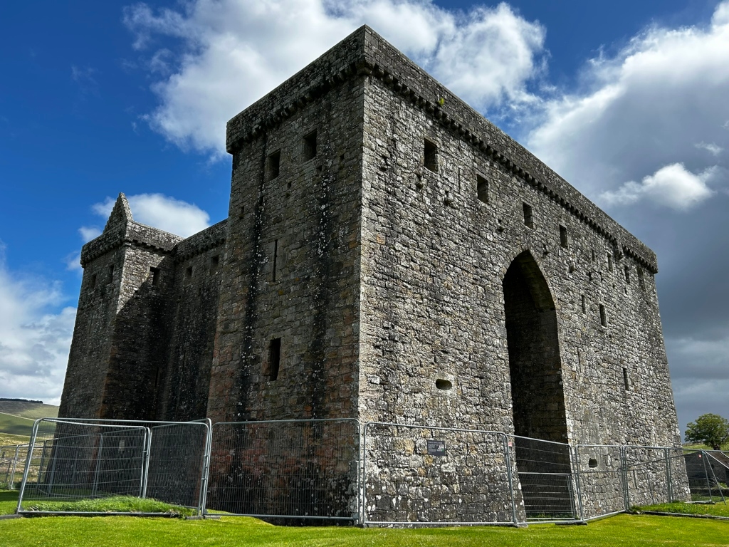 Low-angle, wide shot of a large, grey stone castle under a partly cloudy sky. The castle is partially surrounded by a grey metal fence. The castle's most prominent feature is a large, arched gateway. The stonework is weathered and shows its age. The scene is peaceful and suggests history and possibly some level of preservation or restoration efforts due to the fencing.