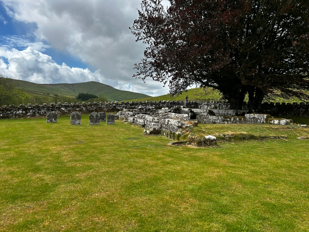 Ruins of a small, ancient structure, possibly a chapel or burial ground, nestled within a grassy field. Several weathered headstones are visible in the foreground, suggesting a cemetery. The stone walls of the structure are partially collapsed, overtaken by moss and grass. A large, dark-leaved tree casts its shadow over the ruins. In the background, rolling green hills under a partly cloudy sky complete the scene, suggesting a remote, rural setting. The overall mood is one of peaceful solitude and the passage of time.