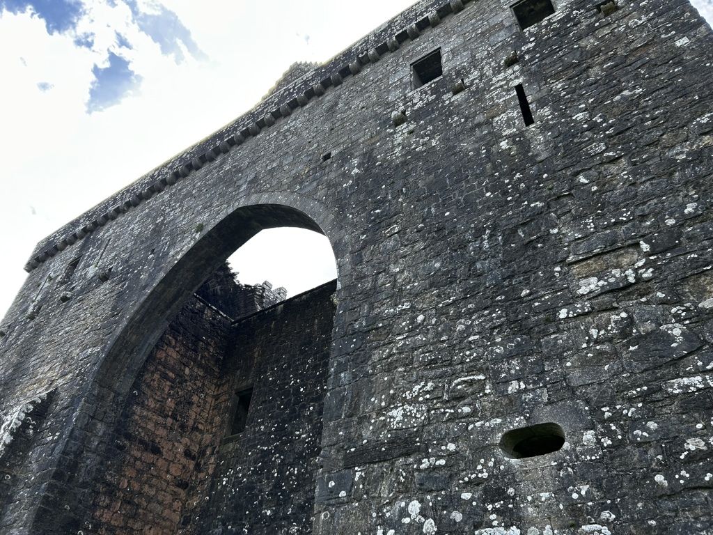 Low-angle view of a section of a stone castle wall. The wall is dark grey stone, heavily textured and showing signs of age and weathering. A large, arched opening dominates the center, revealing a glimpse of the interior structure. Smaller, rectangular openings, possibly windows or arrow slits, are visible higher up on the wall.