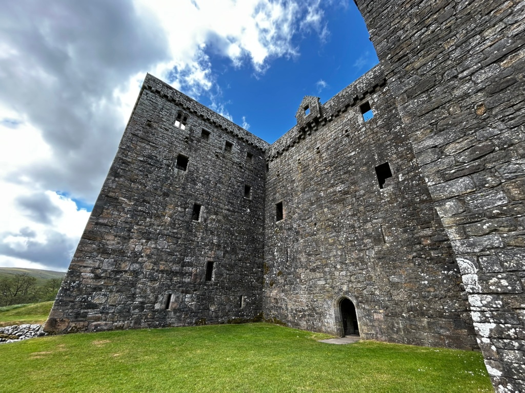 Low-angle view of a corner of a stone castle ruin. The walls are made of rough, grey stones, and there are several small, dark window openings visible. A doorway is located near the base of one of the walls. The foreground shows a patch of green grass. The background includes a partly cloudy blue sky.