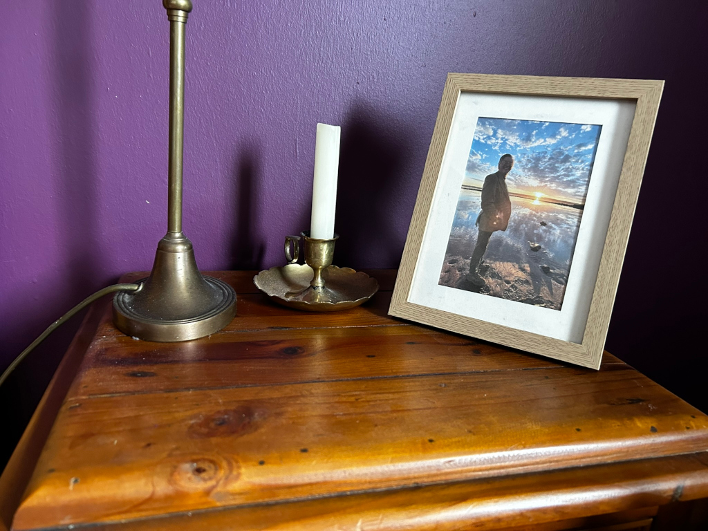 Wooden bedside table against a purple wall. On the table, there is a brass table lamp, a brass candle holder with a white candle, and a framed photograph. The photograph depicts a person silhouetted against a sunset over a body of water.