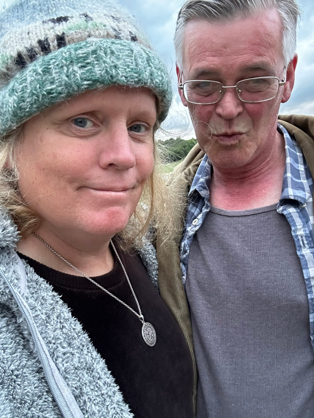 Close-up selfie of a middle-aged couple. The woman is wearing a teal knitted hat and a fluffy grey jacket, and a silver necklace with a circular pendant. The man is wearing glasses and a corduroy jacket over a grey ribbed undershirt. They appear to be outdoors, possibly in a field, with a slightly overcast sky visible in the background. The woman's expression is neutral to slightly amused, while the man's is more puckered and playful. The overall impression is one of casual intimacy and comfortable companionship.