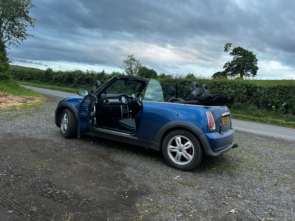 Blue Mini Cooper convertible parked on a gravel road in the countryside. The driver's side door is open, and the car appears to be stopped for a rest stop or scenic view. The sky is overcast with dark clouds, and the overall mood is somewhat serene yet dramatic due to the weather. The rural setting contributes to a sense of peacefulness and escape.