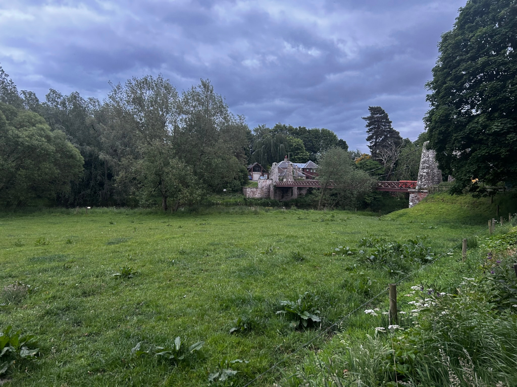 Pastoral scene dominated by a lush green meadow in the foreground. Beyond the meadow, a small, rustic red bridge spans a narrow waterway, leading to a stone building nestled amongst trees. The sky is overcast, giving a somewhat moody atmosphere to the scene.