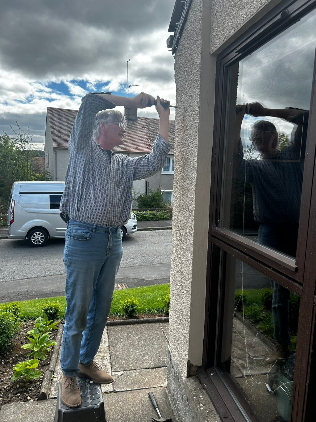 Charlie using a screwdriver to fix something on the exterior wall of a house. He's wearing a plaid shirt, jeans, and boots. He appears to be working on the wall next to a window. The background shows a residential street.