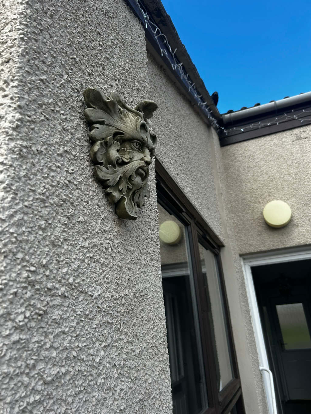 Green Man sculpture affixed to a textured exterior wall of a building.  The Green Man's face is partially obscured by foliage sculpted into the design. The building features a dark brown window and a section of exterior wall with a light fixture. A portion of a second wall and a doorway are also visible. The sky is visible and clear blue. The overall impression is one of a subtle, decorative accent on the building's exterior.