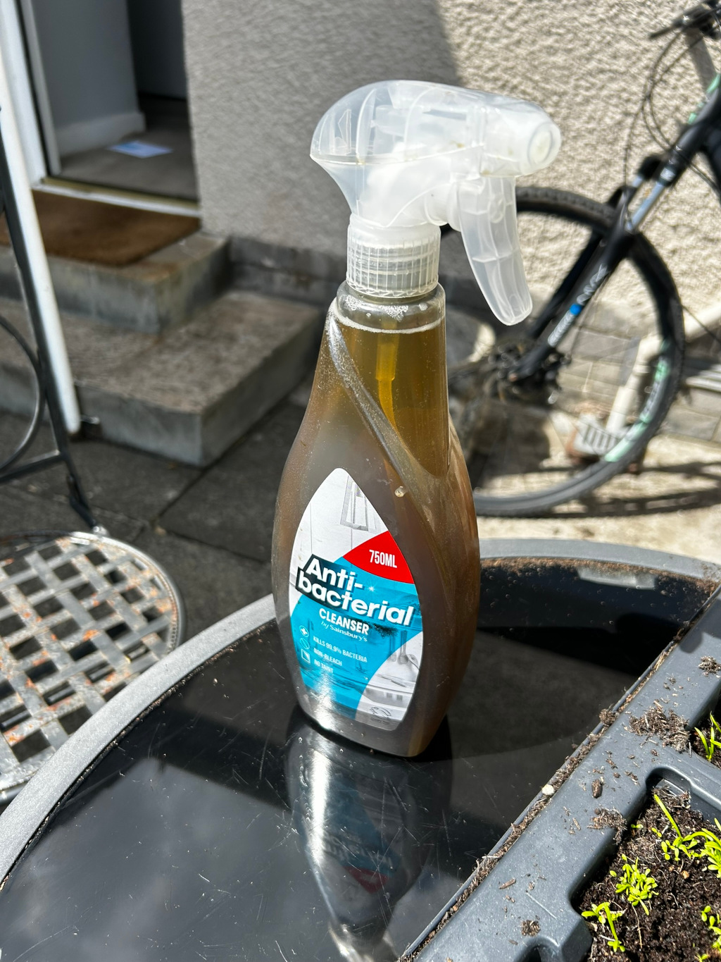 Bottle of Sainsbury's Anti-bacterial Cleanser sitting on a dark-coloured surface outdoors. The bottle is amber-coloured and appears to be partially used, with a visible residue inside. The background shows a portion of a house, steps, and a bicycle. A small garden planter is also partially visible in the bottom right corner.