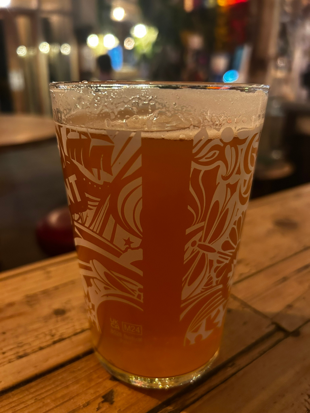 Pint glass of amber-coloured beer. The glass is decorated with a stylised, abstract design in white. The background is blurry but suggests a pub or bar setting. The focus is sharply on the beer glass itself.