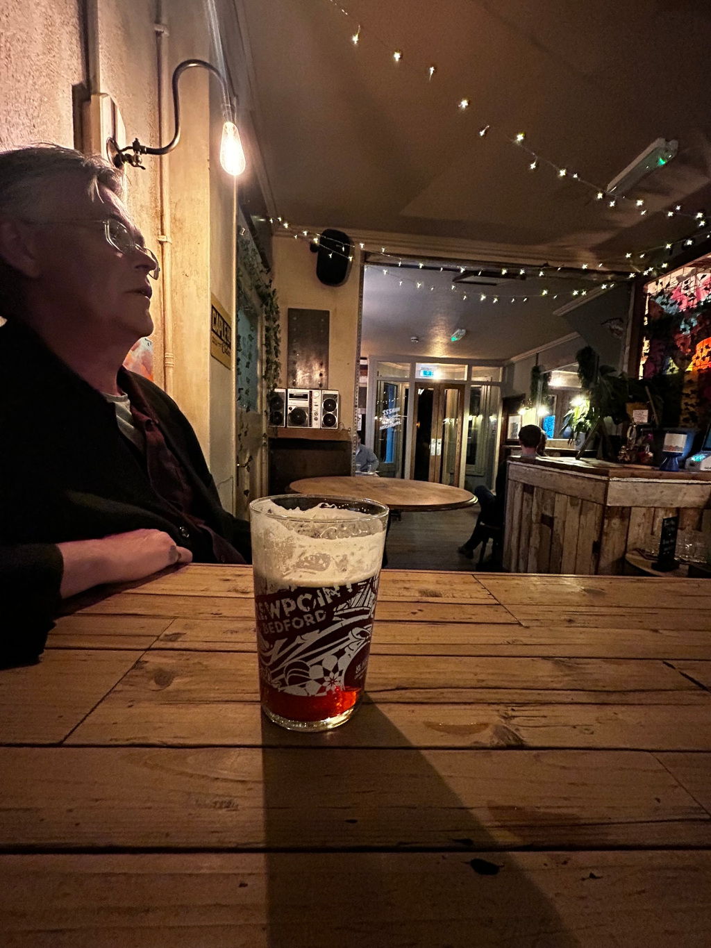 Dimly lit pub interior. Charlie, with grey hair sits at a wooden table with a pint of beer in front of him. The pub has string lights, a rustic aesthetic, and other patrons are visible in the background. The overall mood is relaxed and somewhat contemplative.