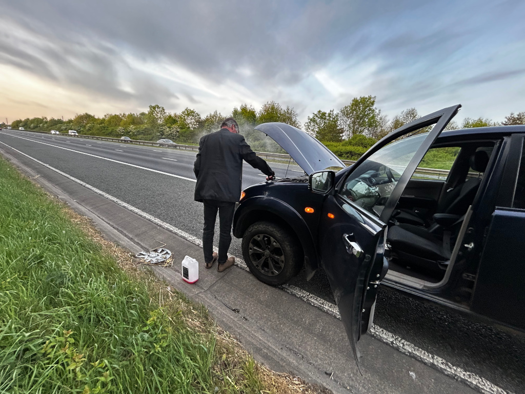 Charlie standing beside his broken-down dark-coloured SUV on the shoulder of a highway. The hood of the vehicle is up, and the passenger side door is open. Charlie appears to be inspecting the engine. The scene is set on a multi-lane highway with other vehicles visible in the distance. The sky is overcast. The overall impression is one of roadside vehicle trouble.