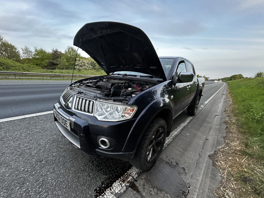 Dark-coloured Mitsubishi L200 pickup truck stopped on the hard shoulder of a highway. The hood is open, suggesting a mechanical failure or breakdown. The truck appears to be leaking some fluid onto the road surface. The setting is a multi-lane highway with green vegetation visible beyond the road. The overall impression is one of a roadside emergency.