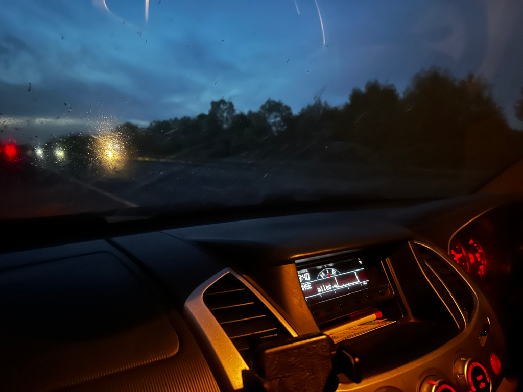 Nighttime shot from inside a our truck. The windshield is misty, and the headlights of other vehicles are visible in the distance. The interior of the car is dimly lit, with the dashboard and instrument panel illuminated. The overall mood is dark, somewhat moody.
