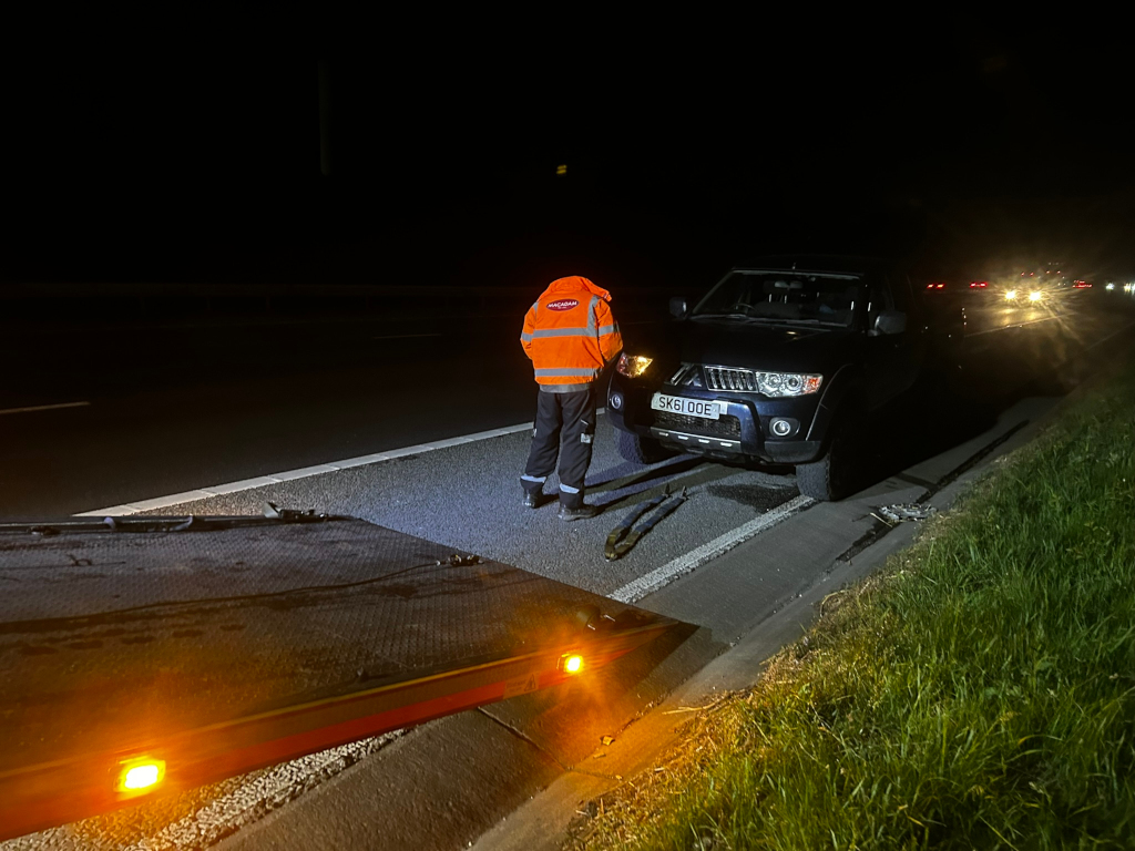 Nighttime scene on a highway. A recovery worker in an orange high-visibility jacket is attending to a dark-colored Mitsubishi L200 pickup truck that appears to be broken down. The truck's license plate is visible. The worker is near the front of the truck, and a flatbed tow truck is partially visible in the foreground. In the background, the headlights of other vehicles on the highway are dimly visible. The overall impression is one of roadside assistance at night.