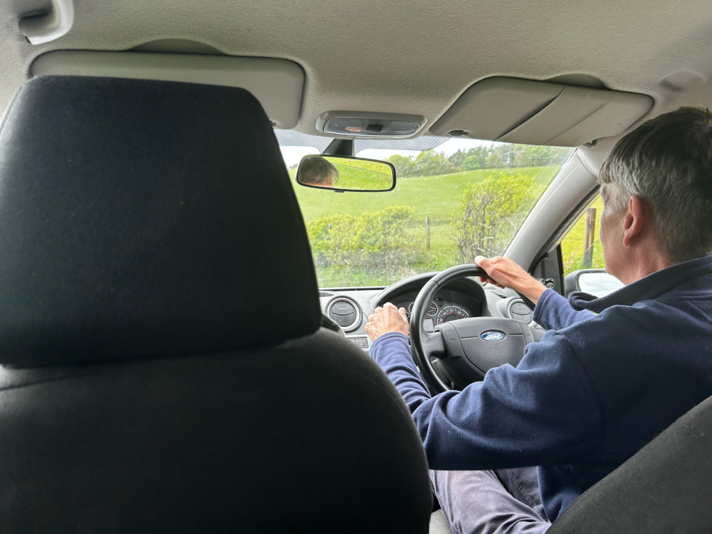 Our neighbour driving a Ford Fiesta. The view is from the back passenger seat, looking forward at the driver. The car is travelling along a rural road, with green fields visible through the windscreen. The overall impression is one of a peaceful, countryside journey.