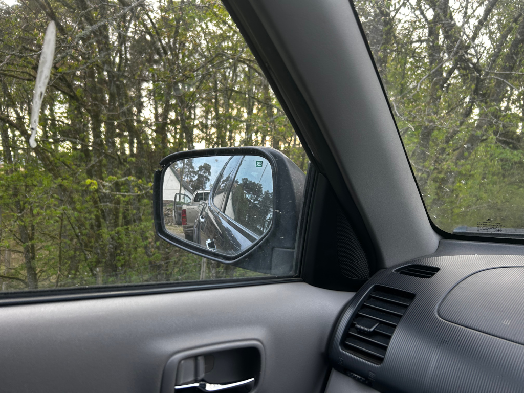 Interior of a car,specifically the passenger's side door and window. The side mirror reflects a view of a wooded area and two other vehicles parked or stopped along a road or driveway. A small building or house is partially visible in the background. The overall scene suggests a rural or wooded setting.