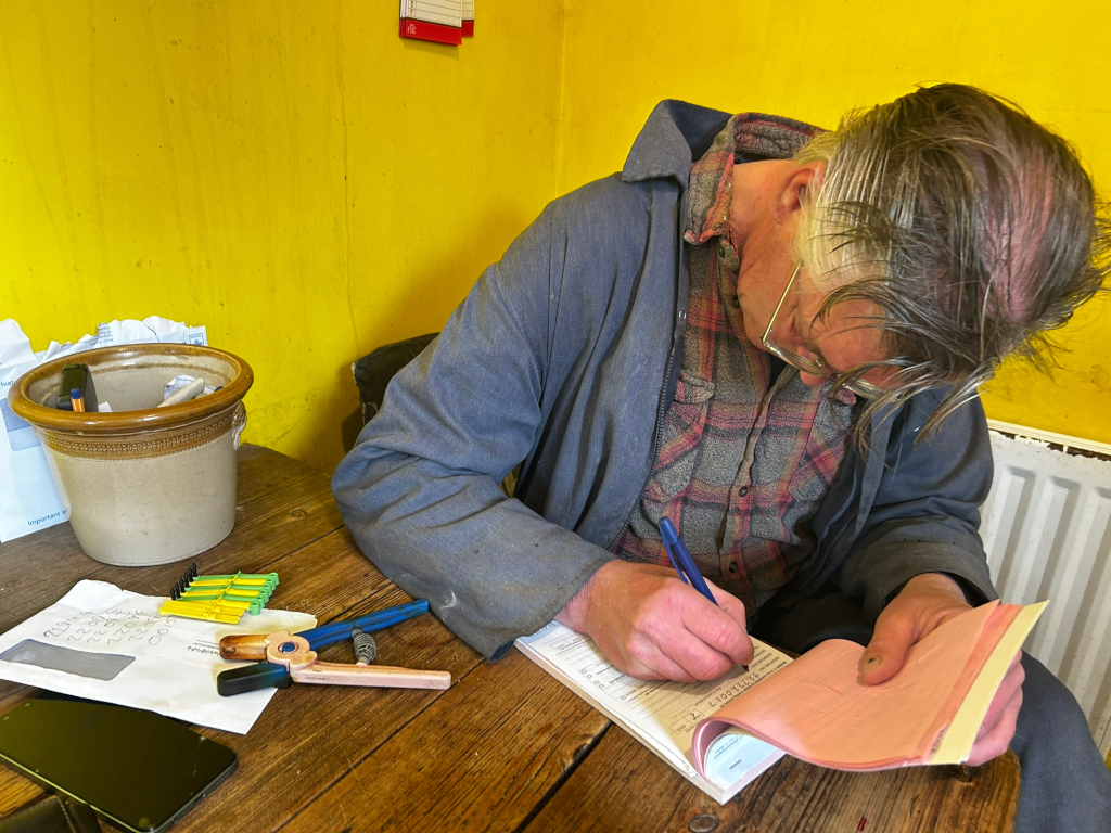 Charlie dressed in a work jacket and plaid shirt, seated at a wooden table. He's hunched over, meticulously filling out a pink invoice or order form with a blue pen. The setting appears to be a small, somewhat cluttered office or workshop, indicated by the yellow walls, a pot containing pens and other small items, and various papers on the table. The overall feeling is one of quiet concentration and possibly routine administrative work.