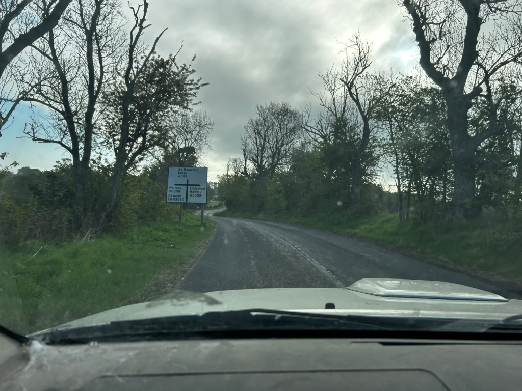 View from inside a car driving down a narrow, winding country road. The road is asphalt and appears somewhat damp. The roadside is lined with lush green grass and mature trees, some bare and others in leaf, suggesting it might be spring. A road sign indicating various directions and distances to different locations is visible on the left. The overall sky is overcast and slightly gloomy. The perspective is from the driver's seat, providing a first-person perspective of the journey.