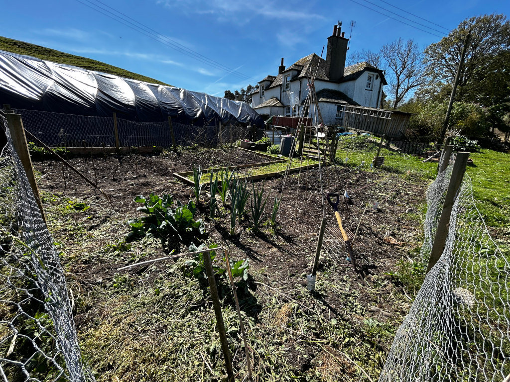 Vegetable garden in the process of being prepared for planting. It's a partially tilled plot of land, enclosed by chicken wire fencing, with some young plants already visible (leeks and possibly cabbages). Gardening tools are lying on the ground. In the background  a stone house is visible. The overall impression is of springtime preparation for gardening, located in a rural setting.